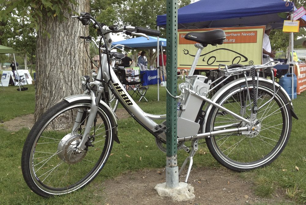 A silver step-through electric bicycle with a front hub motor and a battery pack mounted behind the seat tube, parked on grass at an outdoor event in Reno, Nevada